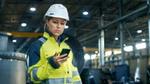 A female factory engineer wearing a hard hat and high-visibility jacket checks her mobile phone