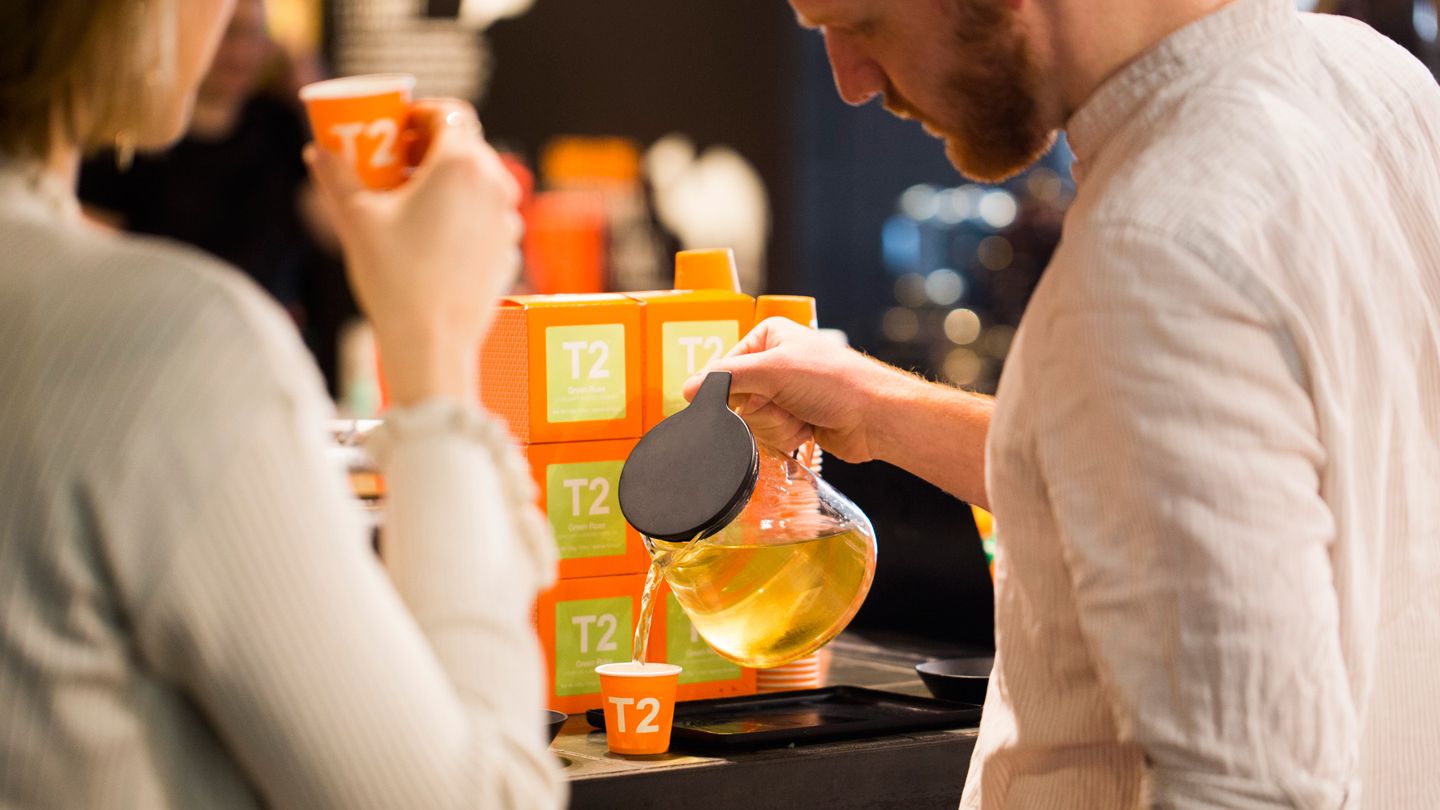 A man pours a glass kettle of tea into a small branded T2 cup in front of a stack of T2 packs. A woman drinks a cup 