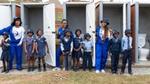 Three women in Domestos-branded blue overalls and several schoolchildren in uniform standing outside a row of toilets.