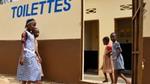 Four young African girls in school uniforms walking into school toilets marked with a painted sign saying ‘toilettes’.