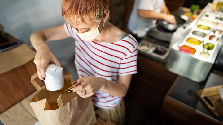 Woman kitchen worker packing food container into a takeaway bag with a chef preparing food in the background.