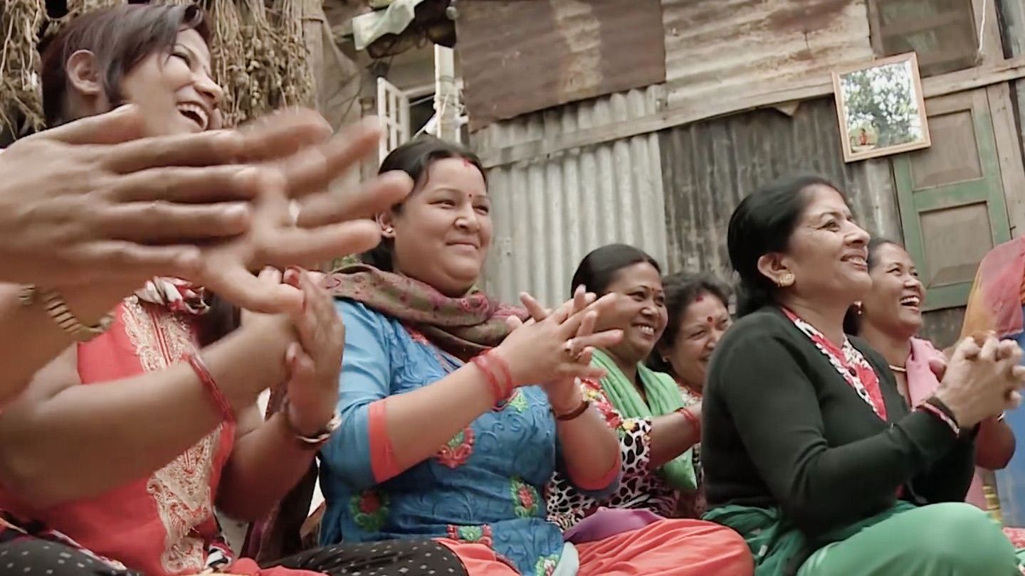 A group of South Asian women in colourful clothes sit on the floor smiling and clapping