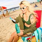 A woman on a beach pours Liquid I.V. powder into a bottle of water to prepare a drink