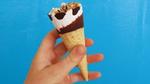 A hand holding a Cornetto ice cream in blue and white packaging. Topping is chocolate and nut. Set on a blue background.