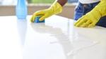 A pair of hands wearing yellow rubber gloves cleaning a white kitchen worktop with a cleaning product
