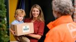 A smiling woman holds her child as she stands at her front door and accepts a parcel from Too Good To Go.