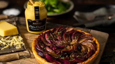 A block of and grated cheese sitting on a grater, a jar of Maille Dijon Mustard, and an onion tart, all on a wooden board.