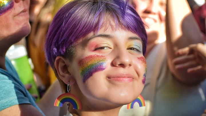 Una chica con cabello morado sonriendo, con banderas del Orgullo pintadas en su rostro