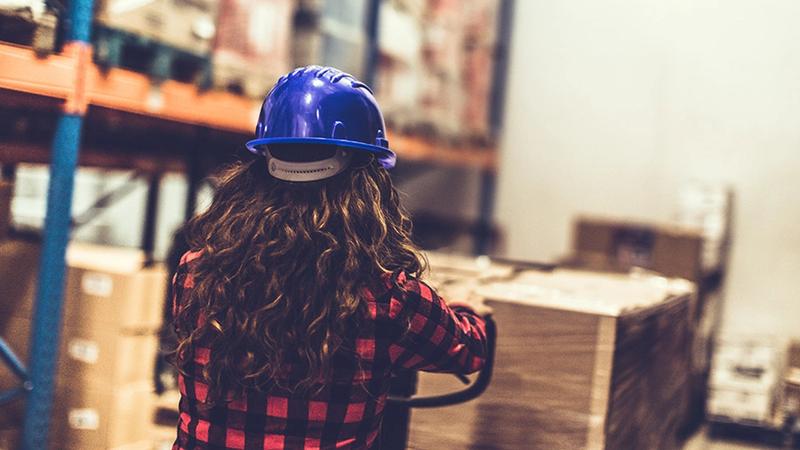 The back of the head of a young woman in a hard hat surveying a warehouse of goods
