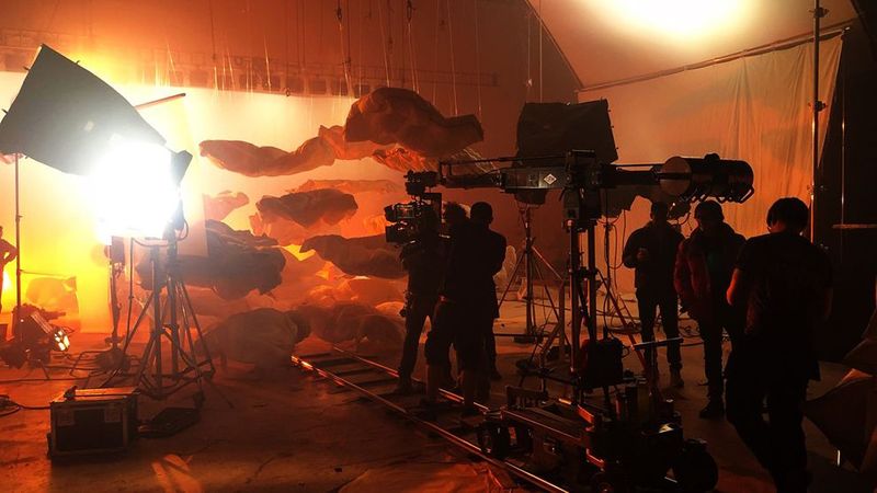 A film crew in a studio is setting up a shot of a large screen with floating grey and orange cut-out clouds.