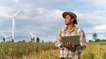 Asian farmer standing in a field, using his laptop to analyse crop data. Wind turbines can be seen in the background.