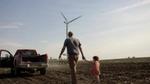 Father and daughter walking hand in hand across soil towards a red truck, wind turbine in the distance