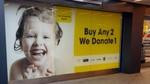 Large window sticker with text 'Buy Any 2 We Donate 1' against a yellow background, seven brand logos, and a photo of a child