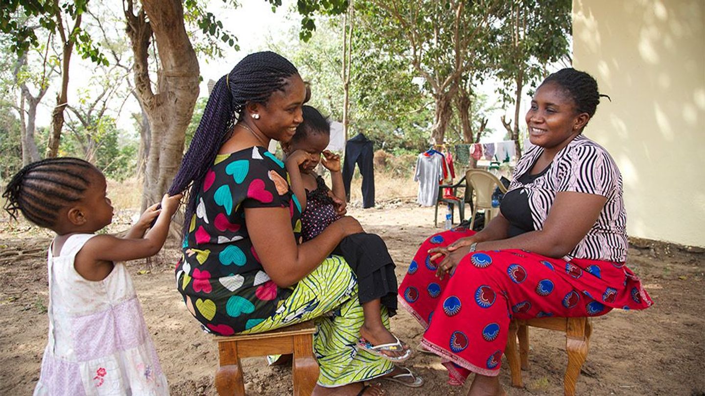 Afrikaanse vrouwen in kleurrijke kleding zitten buiten lachend tegenover elkaar.