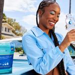 Woman in a blue shirt standing against a blue car, drinking a bottle of Liquid I.V.