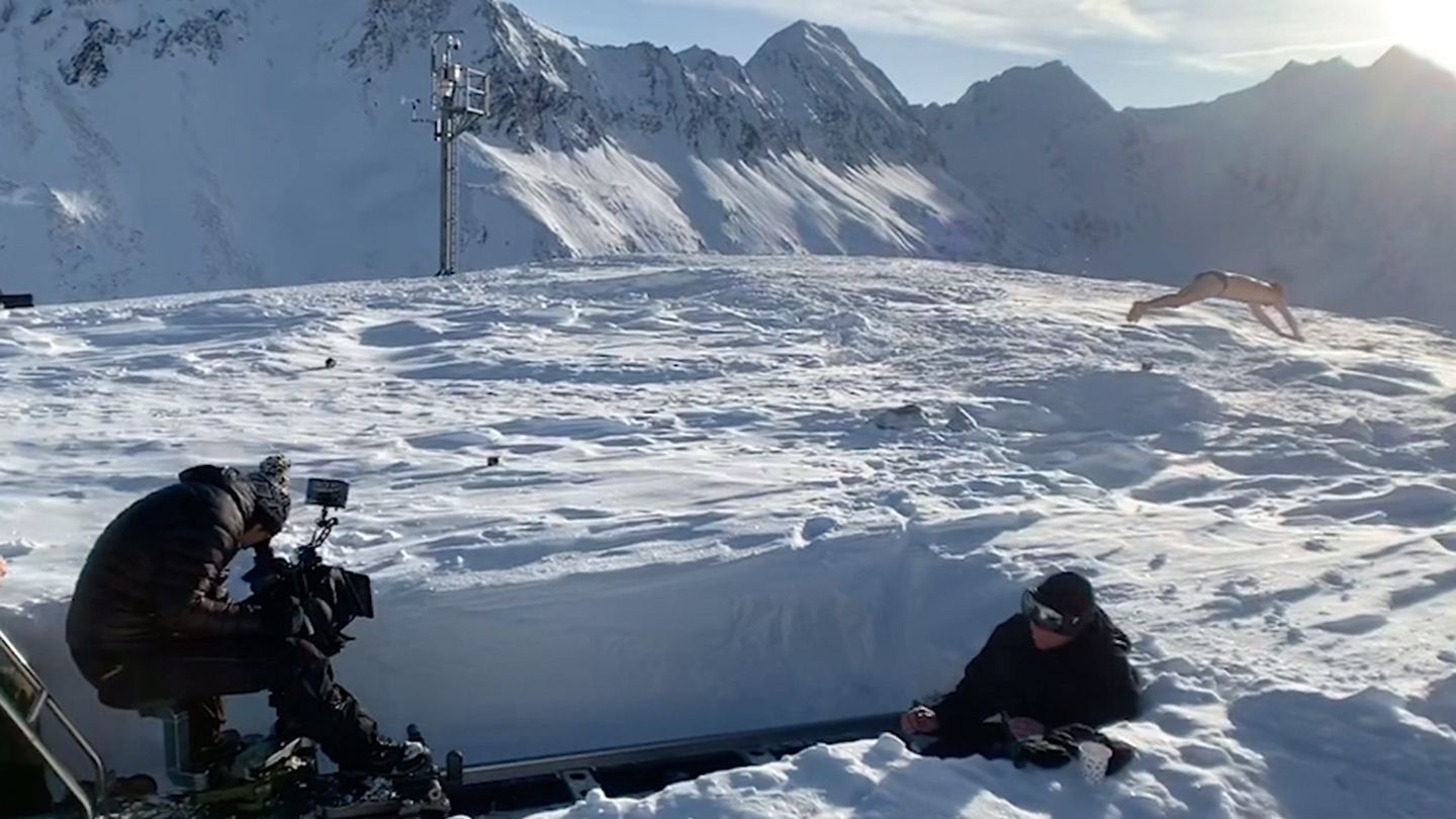 On a sunny snow field surrounded by mountains  a photo crew ski wear is photographing a man doing press-ups in boxer shorts