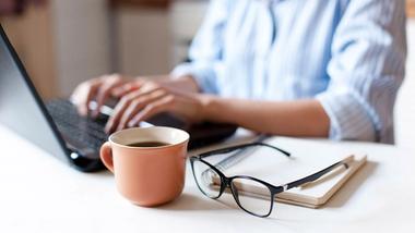 a close up of hands at a laptop with a pair of glasses and a cup of coffee in the foreground