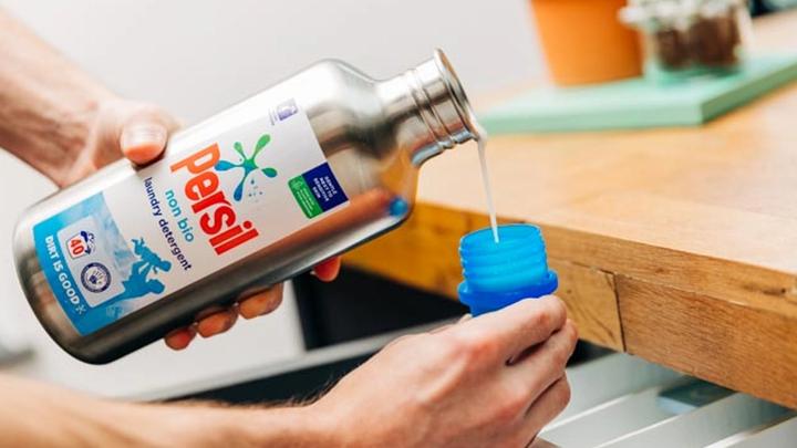  Image shows hands holding a Persil aluminium refill bottle pouring out detergent into a plastic lid]