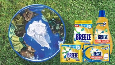 A laundry tub with green, brown and white clothes is placed on top of outdoor grass. Next to it are four Breeze products.