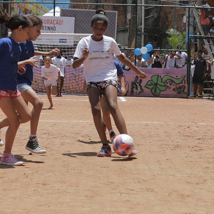 Girls playing football on an inner-city dirt pitch surrounded by fencing, being watched by a small group of people.