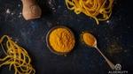 A small bowl of orange microalgae powder on a blue-grey table with two tangles of thick pasta on the side.