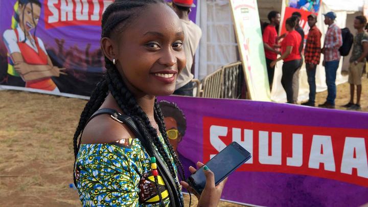 Young woman smiling at the camera, holding a mobile phone, in front of a Shujazz banner at an outdoor event. 