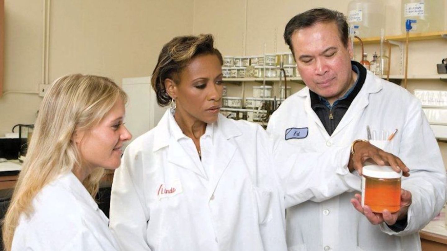 Three people in white lab coats standing in a science lab look closely at a small jar full of a chemical substance.
