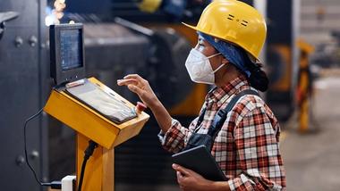 A female factory engineer in a hard hat and mask checks a control panel