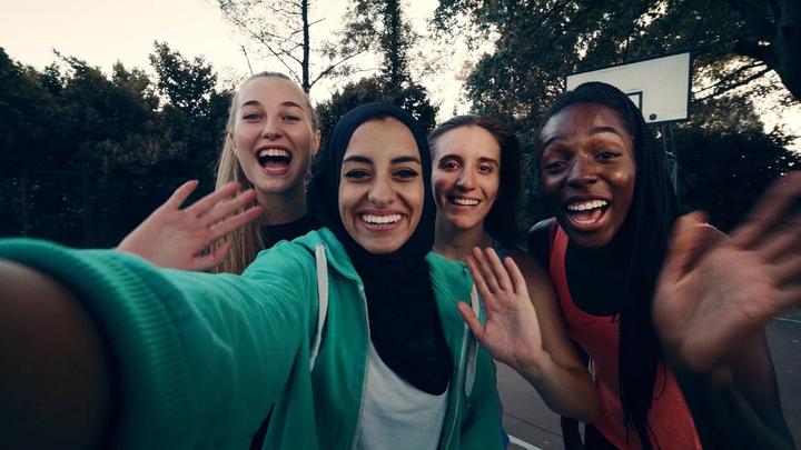 Four women of different races and religion smile and wave to the camera