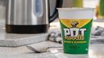 A Chicken & Mushroom Pot Noodle in a paper pot stands on a kitchen counter next to a kettle and a fork. 