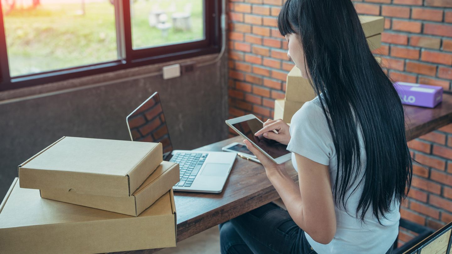 Asian woman at home online shopping. A laptop, an iPad and parcels surround her