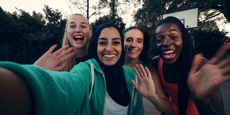 Four women of different races and religion smile and wave to the camera