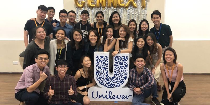 A group of smiling GEN-NEXT interns pose for the camera around a Unilever logo / --
