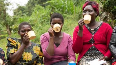 Three women drink tea from tin mugs during a break in a farmer field school training session