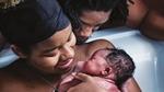A mother happily cradles her newborn baby in a birthing pool. A man at her side looks on proudly.