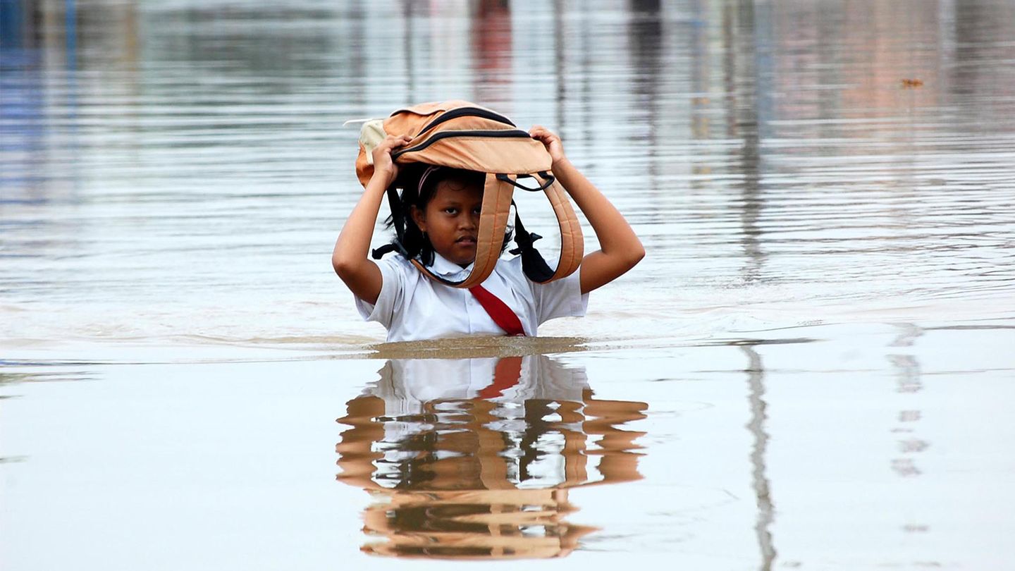 Schoolgirl wading through flood waters with her school bag above her head 