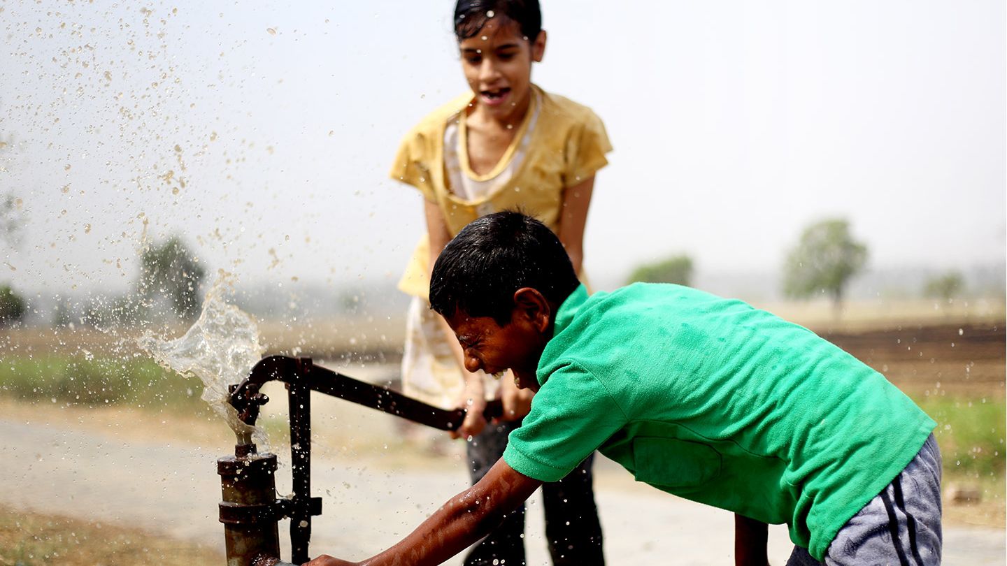 Two children playing and drinking water by an outdoor water tap