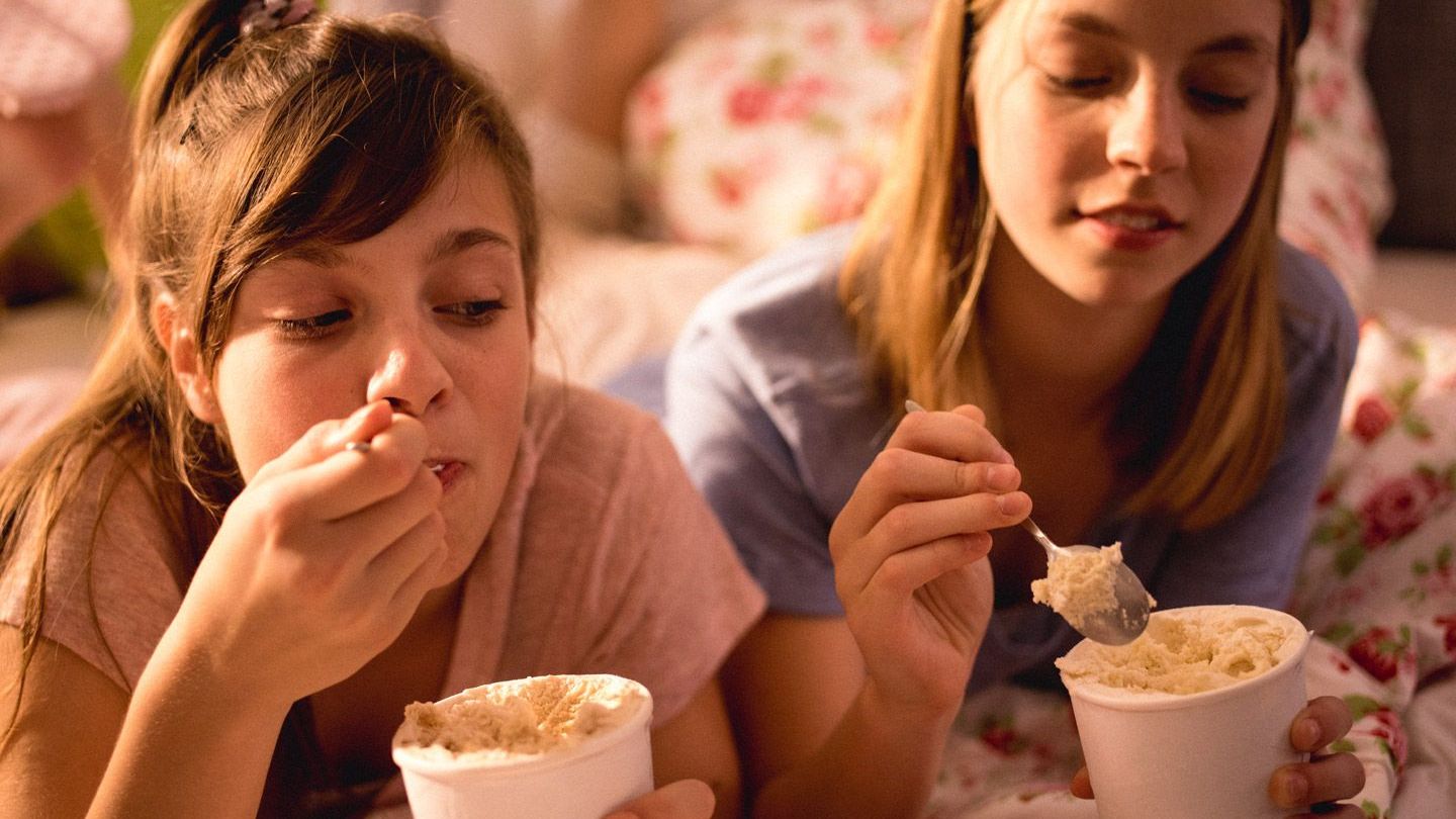 Two teenage girls lie on a bed eating big tubs of ice cream