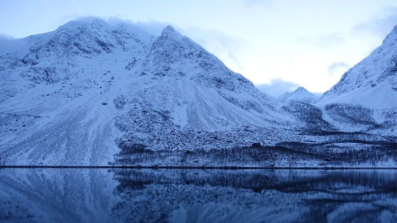 Ice capped mountains sit behind a lake. The reflection of the mountains is mirrored in the lake