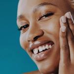 A woman with short, bleached hair applies skin cream to her cheek. She smiles at the camera.