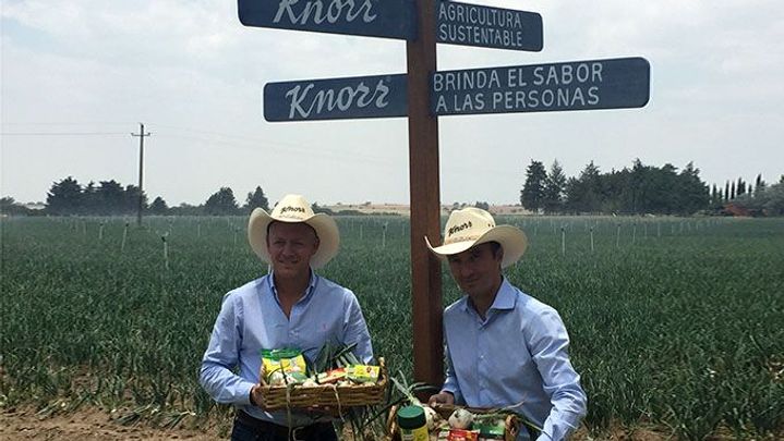 Dos personas con un campo detrás y carteles de Knorr