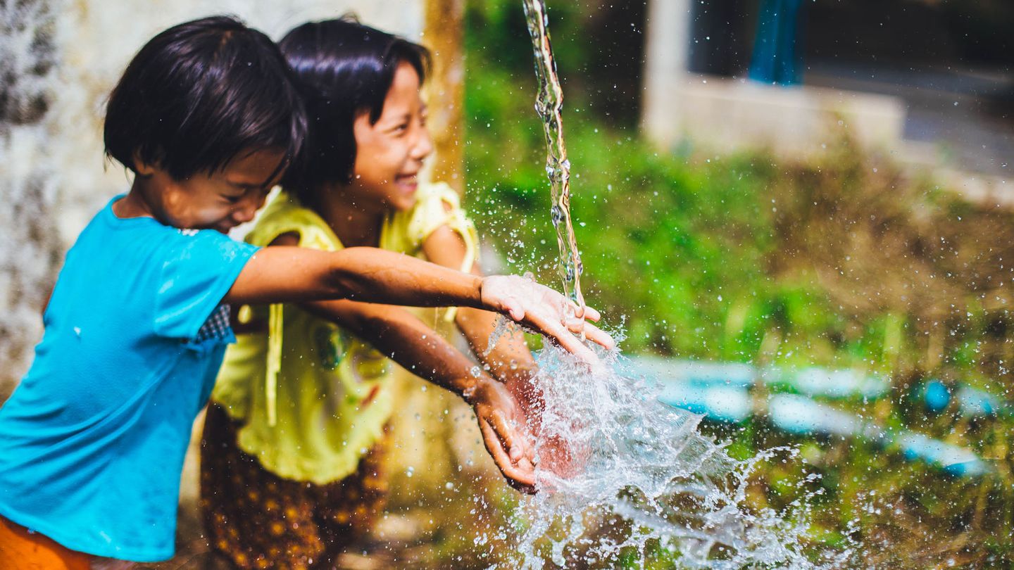 Young boy and girl smiling, holding their hands under the spray of water from outside tap.