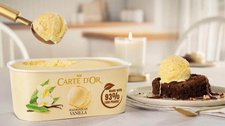 Tub of vanilla Carte D’Or ice cream on table and scoop on top of chocolate dessert.