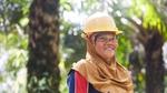A woman in a headscarf and yellow hard hat stands in focus smiling with palm trees behind her