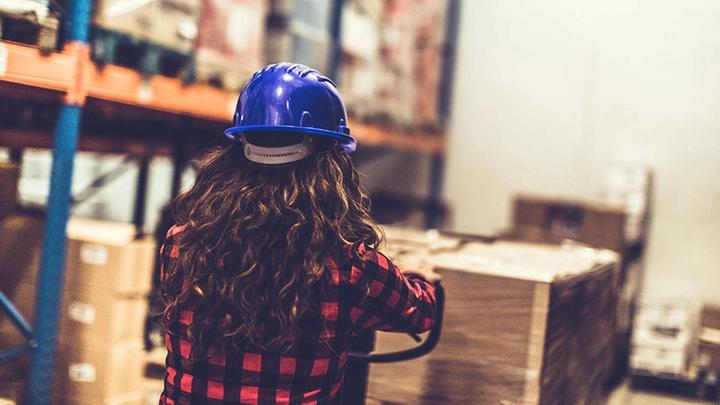The back of the head of a young woman in a hard hat surveying a warehouse of goods