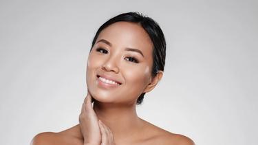A Filipina woman is smiling at the camera while gently placing her hands near her cheek and neck.