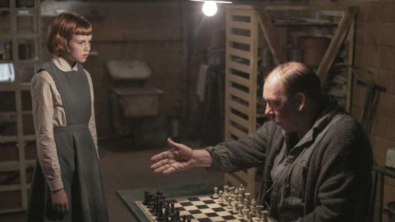Mr Shaibel pointing at Beth to sit down in front of the chess board. 
