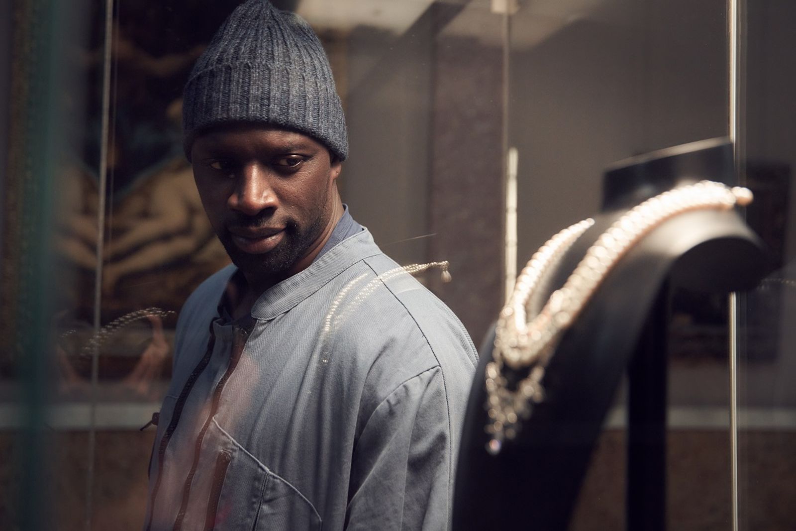 Assane played by Omar Sy is staring into a glass cabinet with jewels in.