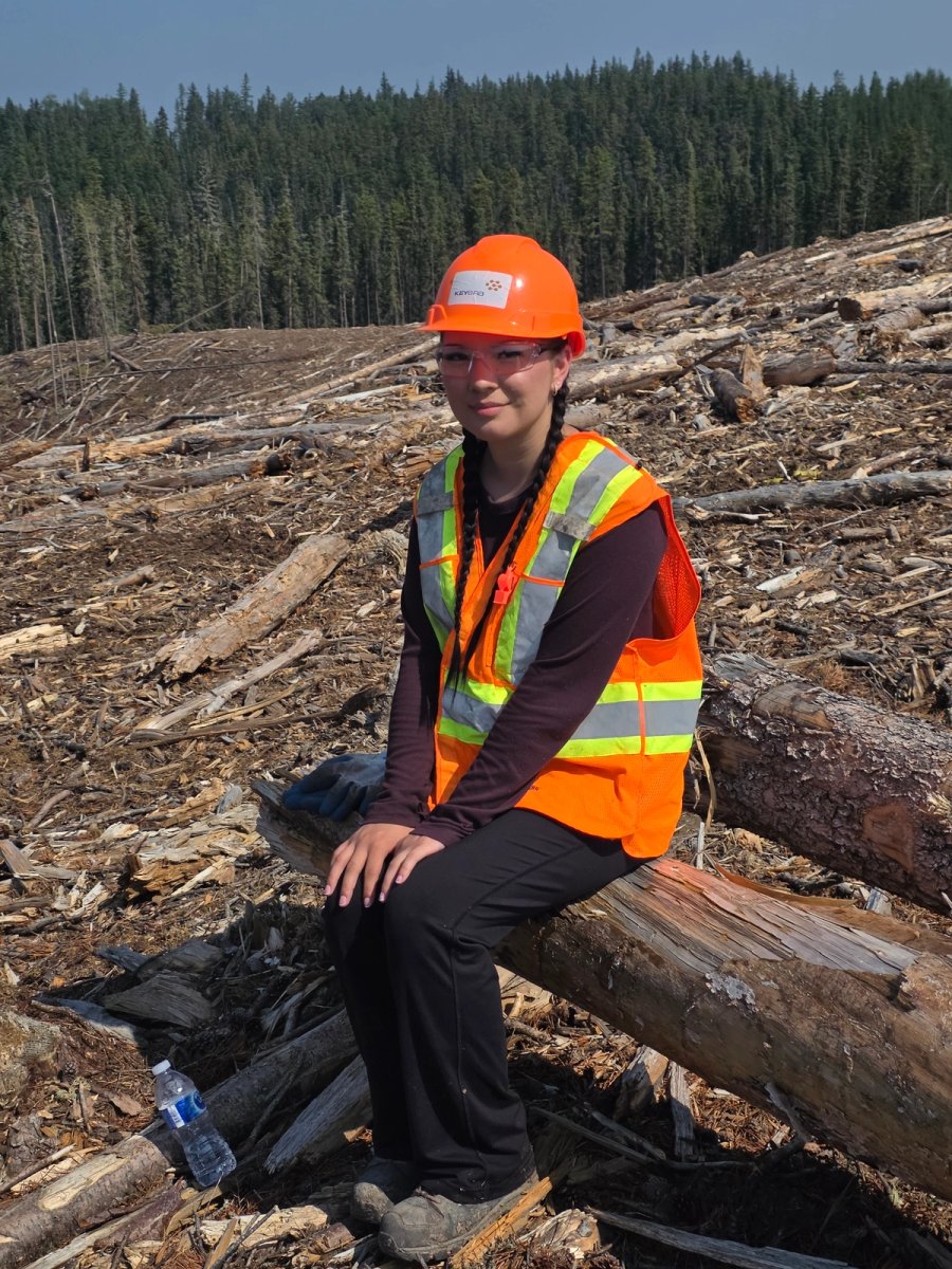 Smiling young woman in a hard hat and safety vest sits on a log in a clear-cut forest, with trees in the background.