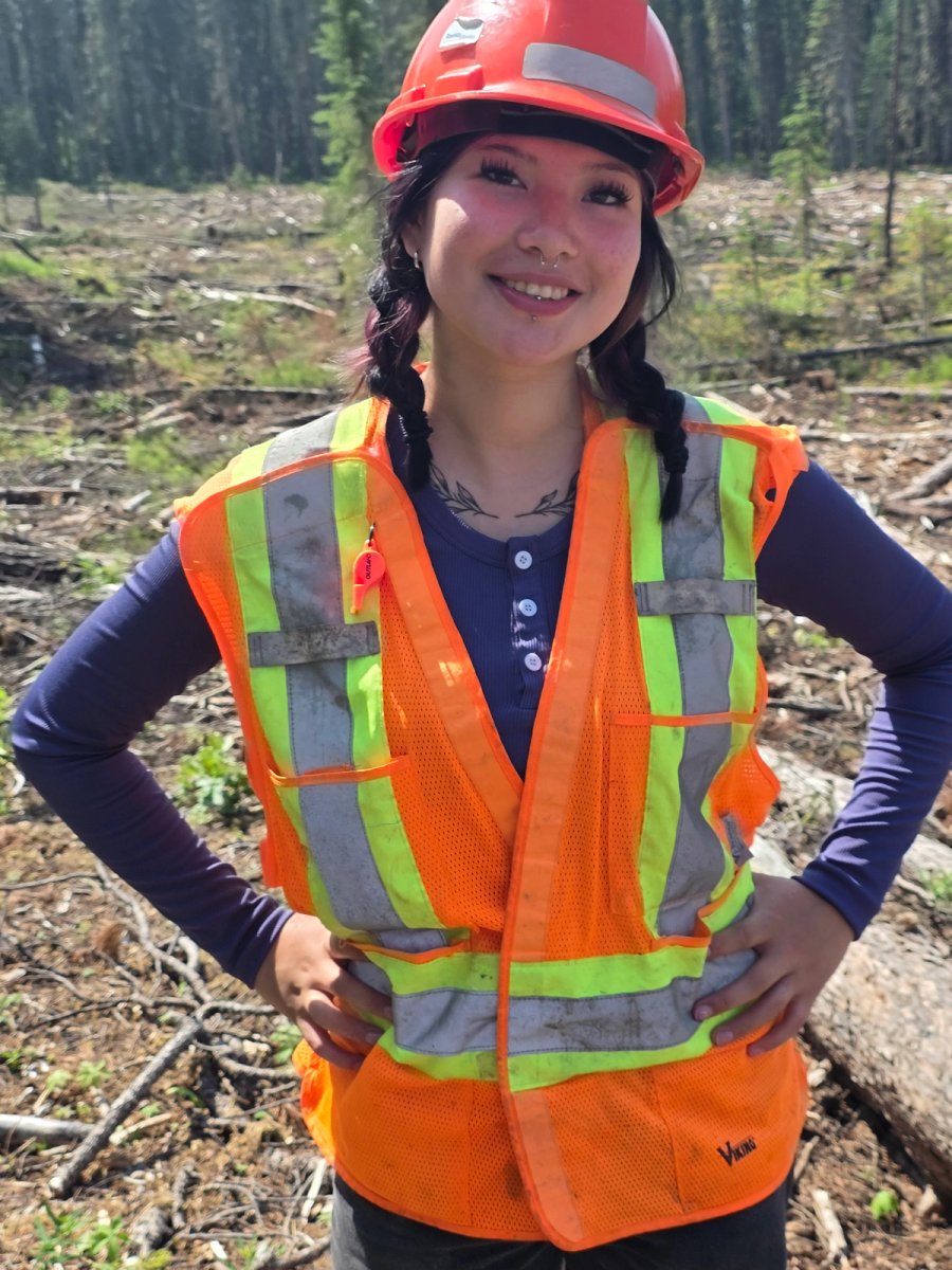 A young woman in an orange hard hat and reflective vest smiles in a cleared forest.